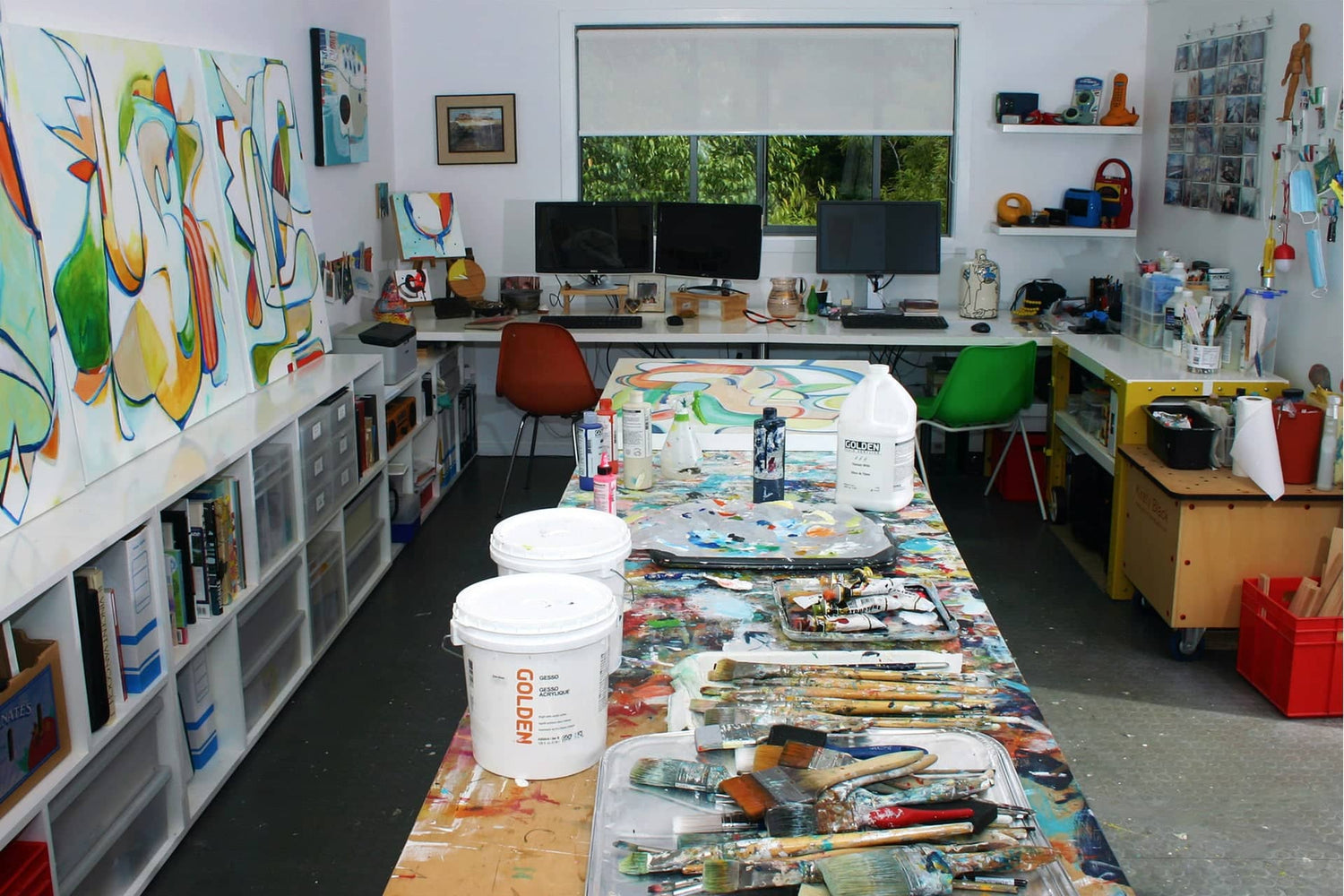 Kirsty Black's art studio in Maraetai, Auckland, showing a large paint-covered work table with brushes and Golden acrylic paints, original abstract paintings on the wall, and shelving filled with art materials and books.