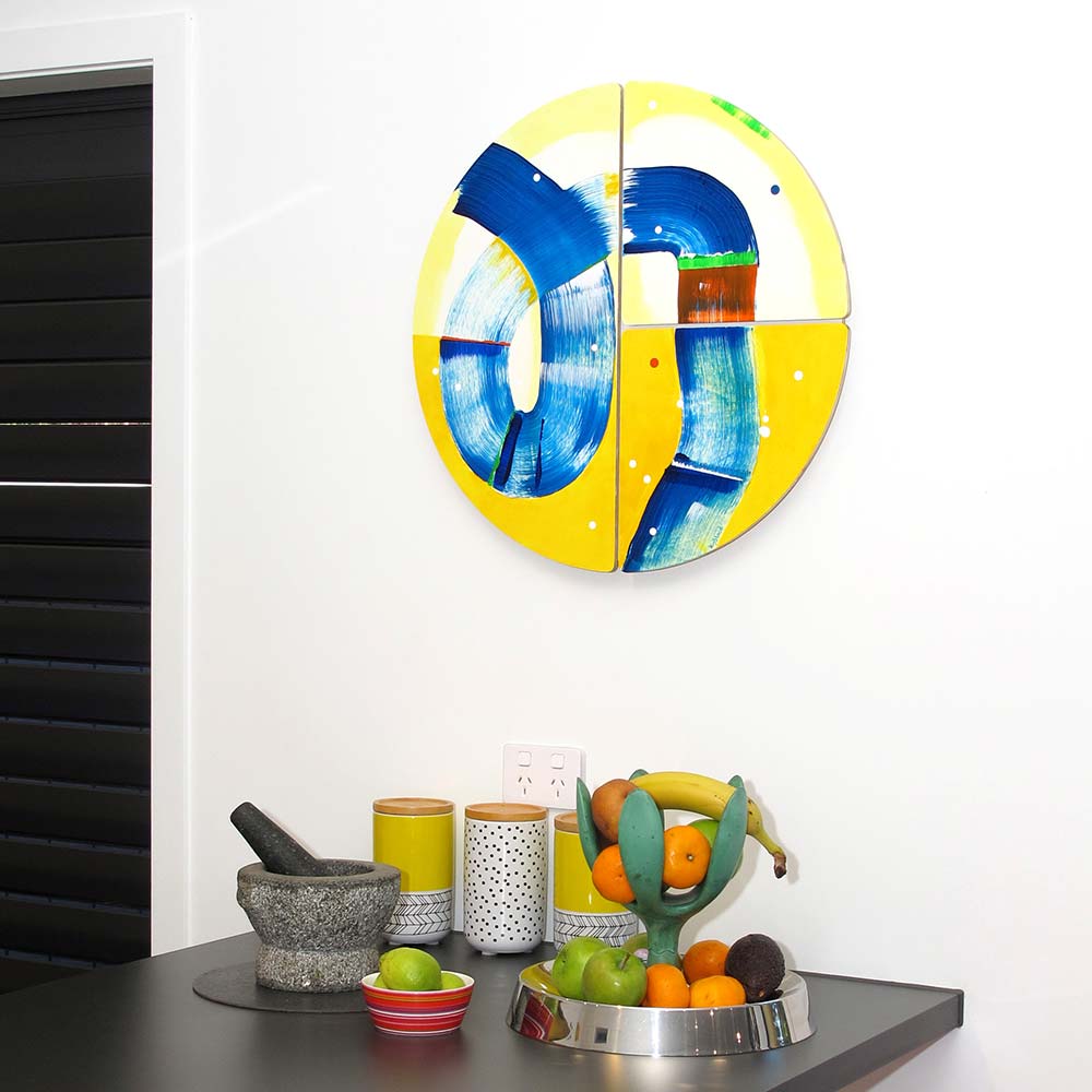 Hippo Water Chute wall sculpture by Kirsty Black, its bold cobalt blue and vivid yellow forms radiating energy above a dark kitchen bench with fruit bowl and canisters in a clean New Zealand interior.