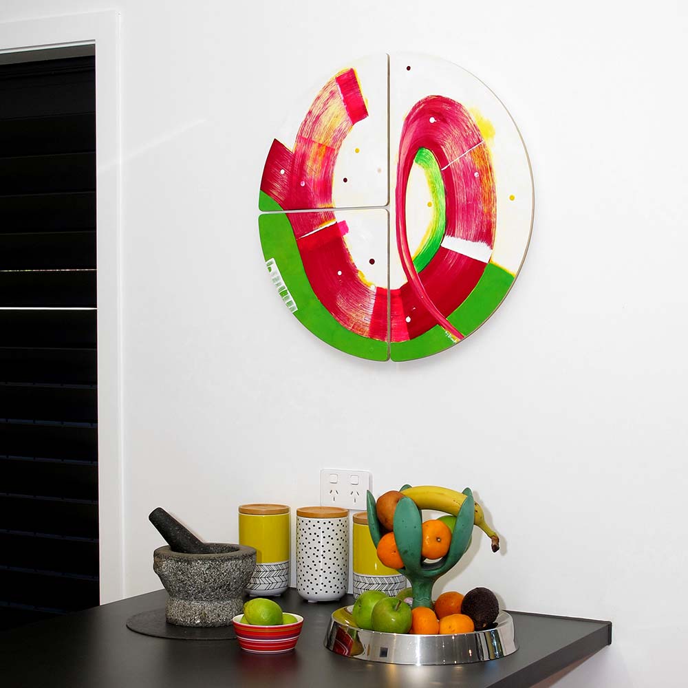 Sausage Dog Wheelie wall sculpture by Kirsty Black, its vivid crimson and green forms popping against a white wall above a dark kitchen bench with fruit bowl and canisters.