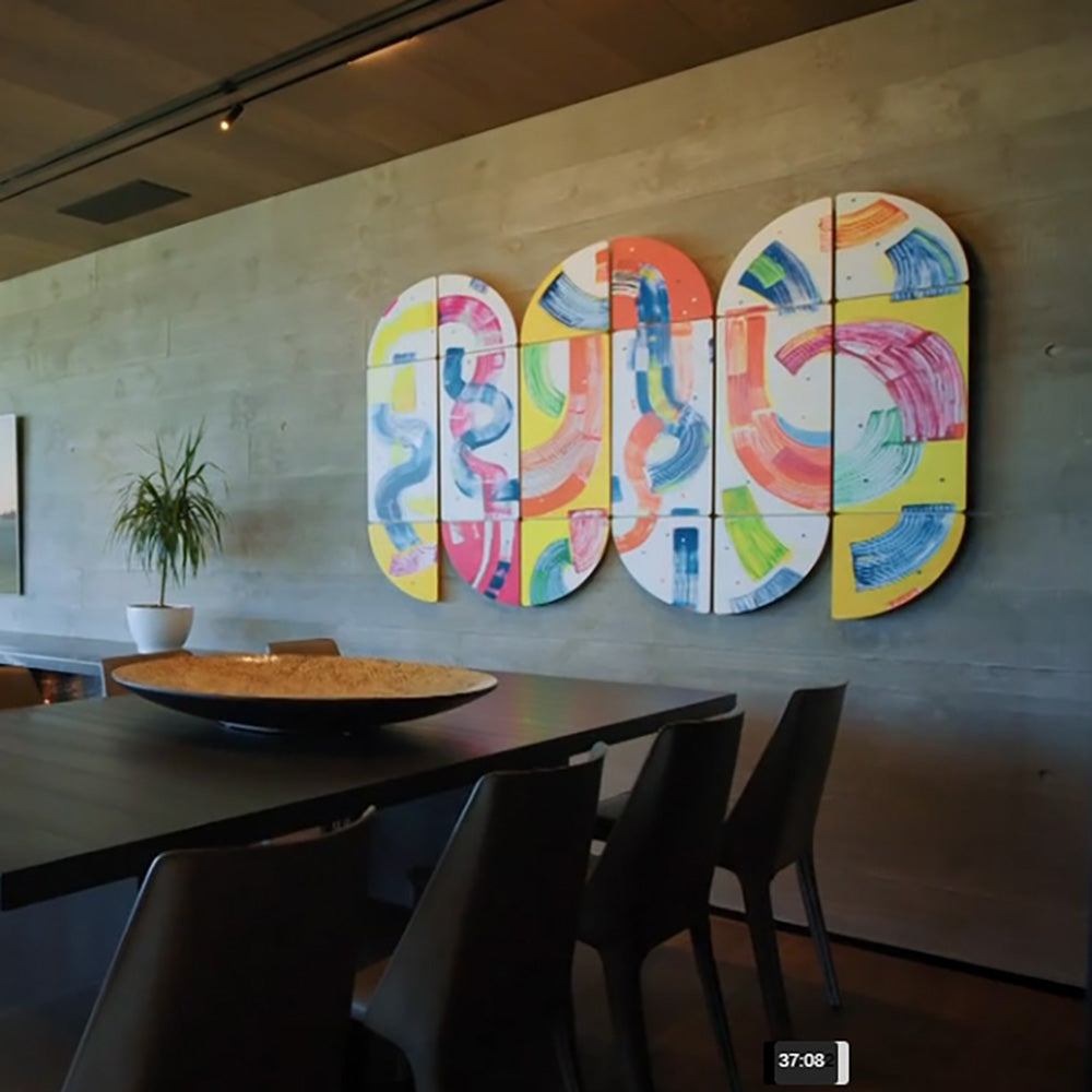Carnival Cavort wall installation by Kirsty Black installed on raw concrete wall in architect-designed dining room featured on Grand Designs New Zealand, comprising multiple shaped panels in vibrant yellow, pink, orange, blue and green above a dark timber dining table